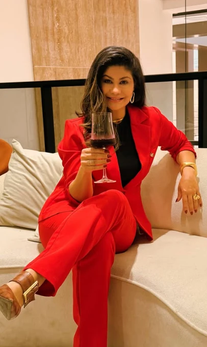 Lucineia Campos sentada em um sofá claro, de terninho vermelho, segurando uma taça e sorrindo.